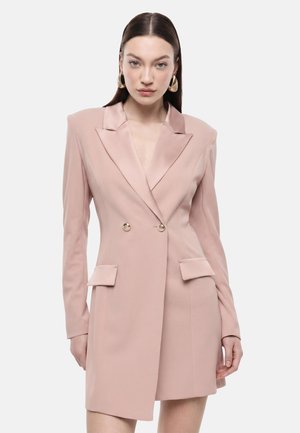 Donna con capelli castani lisci che indossa orecchini a cerchio dorati e un vestito blazer doppiopetto rosa chiaro con tasche a patta.