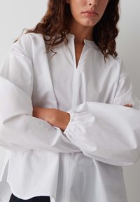 Weiße Baumwollbluse mit hohem Kragen, langen Puffärmeln und einem seitlichen Schlitzsaum. Verfügt über eine Vorderleiste und eine glatte Textur.