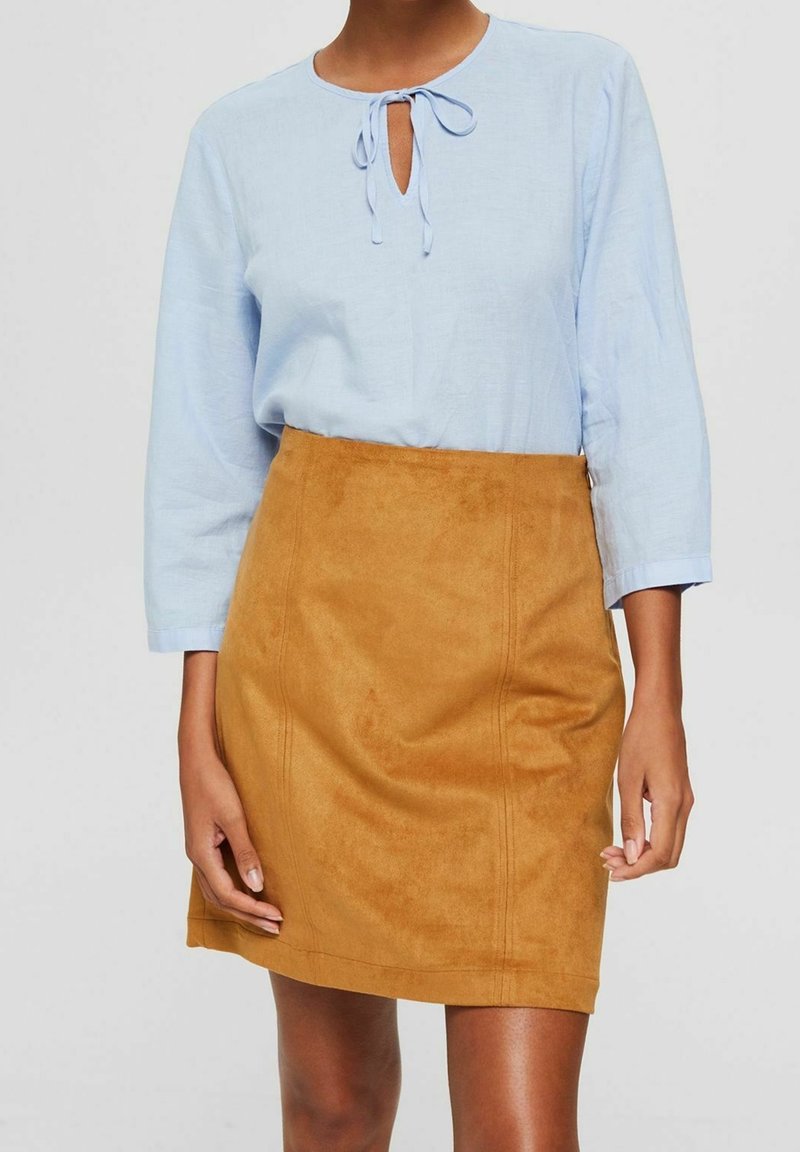 Femme portant un chemisier bleu clair à col noué et une mini-jupe en daim couleur camel, debout devant un fond uni.