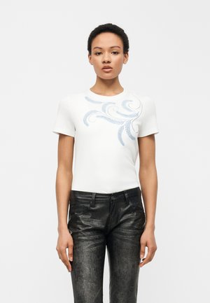 Frau trägt ein weißes kurzärmeliges Oberteil mit blauen Blattmustern auf der Brust und schwarze, strukturierte Lederhosen, steht vor einem einfarbigen Hintergrund.