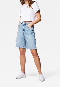 Donna che indossa una maglietta bianca corta, pantaloni corti in denim bleu chiaro a vita alta con orli sfrangiati e sneakers bianche, in piedi con una mano nella tasca.