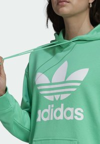 Zelená mikina s kapucí s velkým bílým logem Adidas se třemi pruhy a ikonickým listovým motivem; obsahuje stahovací kapuci a žebrované manžety.