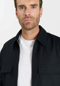 Zwarte jas met ritssluiting en kraag, met twee voorzakken, gedragen over een wit T-shirt. Soepele textuur en minimalistisch ontwerp.