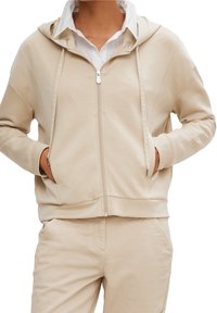 Beige zip-up hoodie met een zachte textuur, zijzakken en een capuchon. Heeft subtiele branding langs de rits en een relaxte pasvorm.