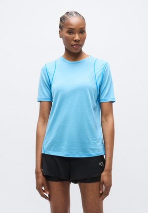 Svetlo modra športna majica s kratkimi rokavi, zaobljenim izrezom in kontrastnim šivanjem. Oblečena skupaj s črnimi kratkimi hlačami, ki imajo na spodnjem robu logotip.