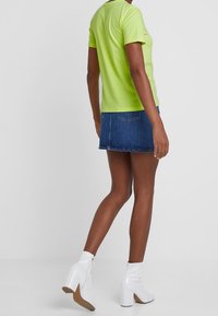 Jasno-limonkowa koszulka z krótkim rękawem w połączeniu z ciemnoniebieską mini spódniczką z dżinsu. Modelka nosi białe botki za kostkę na grubym obcasie.