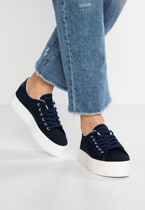 Person iført blå jeans med flossede kanter og marineblå snørestøvler med plateau på hvidt gulv.