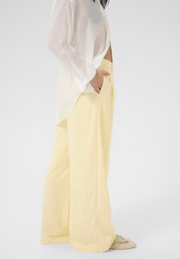 Pantaloni larghi giallo chiaro realizzati in tessuto morbido, con tasche laterali. Abbinati a una camicia bianca trasparente e leggera.
