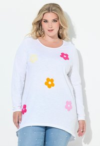 Blondýnka v bílém topu s dlouhým rukávem, na kterém jsou barevné aplikace květin, a v světle modrých džínách zdobených perlami.