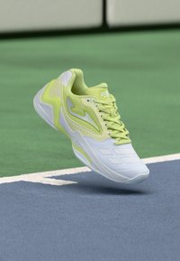 Αθλητικό παπούτσι ανοιχτού πράσινου και λευκού χρώματος με πλέγμα και συνθετικά πάνελ, που αιωρείται πάνω από την επιφάνεια γηπέδου τένις σε μπλε και πράσινο χρώμα.