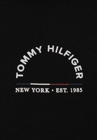 Tela negra que presenta un logotipo de texto arqueado en blanco que dice "TOMMY HILFIGER" sobre "NUEVA YORK • FUNDADA EN 1985" con rayas de acento rojas y azules debajo.