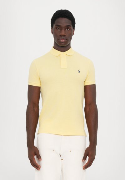 Camisa polo amarilla de manga corta, con tejido texturizado, cuello y un pequeño logo en navy en el lado izquierdo del pecho, combinada con pantalones de color claro.