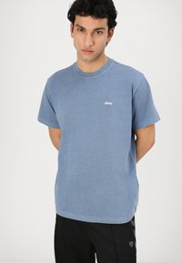 Giovane uomo con capelli ricci che indossa una maglietta blu "obey" e pantaloni neri, in piedi con le mani dietro la schiena davanti a uno sfondo semplice.