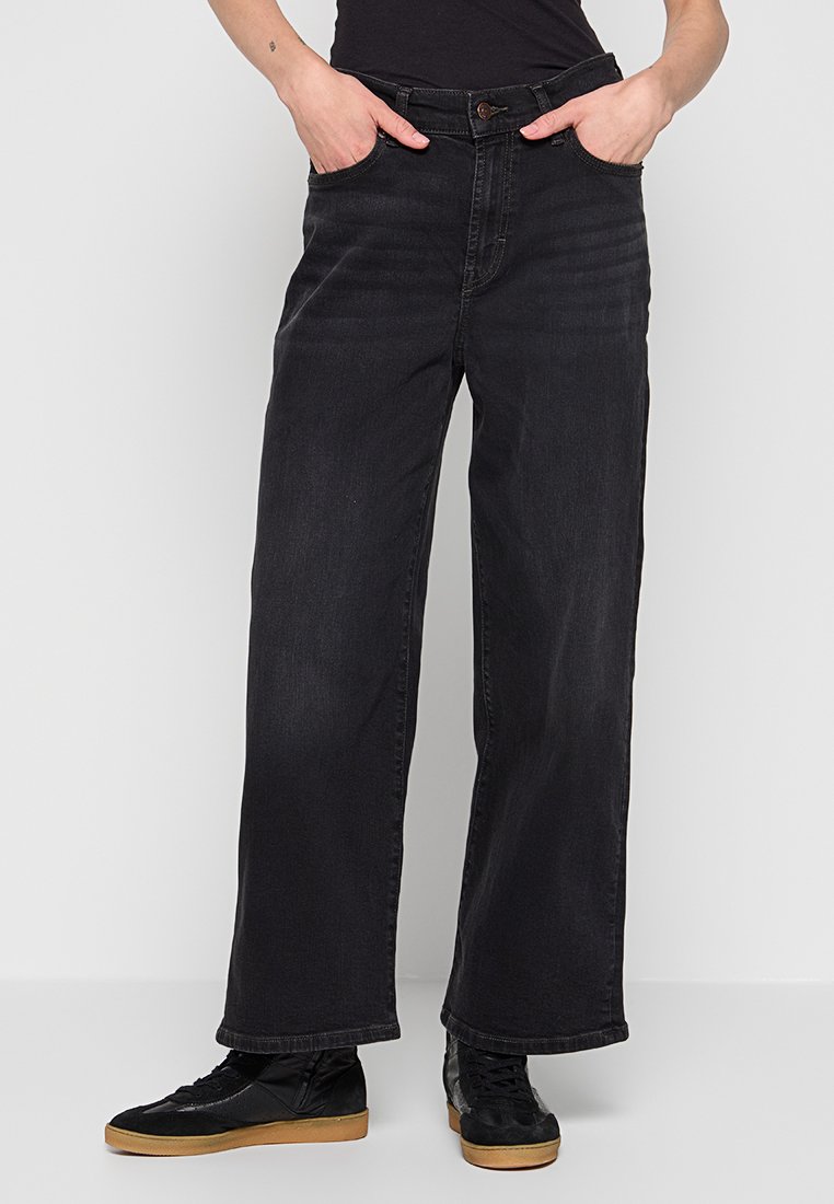 Person in weit geschnittenen schwarzen Jeans mit den Händen in den Taschen und schwarzen Sneakers mit Gummisohle, der vor einem schlichten hellen Hintergrund steht.