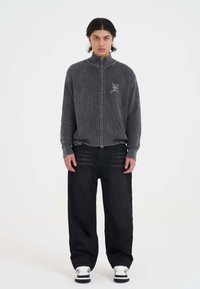 Young man with dark hair wearing a ribbed grey zip-up sweater, loose black jeans, and white sneakers standing against a plain white background.