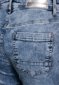 Jeans de mezclilla azul claro con un bolsillo trasero que presenta un detalle de costura roja y un parche de logo plateado en la cinturilla. Textura suave.