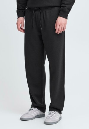 Black sweatpants made of soft fabric, featuring an elastic waistband with a drawstring, side pockets, and relaxed fit. Paired with gray sneakers.