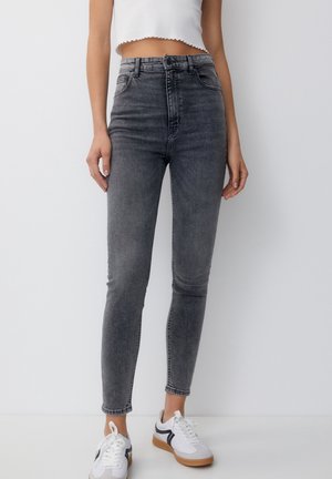 Woman wearing high-waisted faded black skinny jeans, white cropped tank top, and white sneakers with black and brown details.
