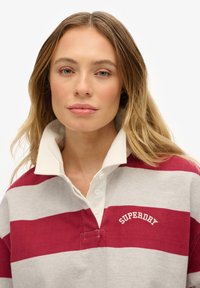 Maglia da rugby a righe rosse e grigie con colletto bianco, dotata di patta con bottoni e logo "Superdry" ricamato sopra il petto.