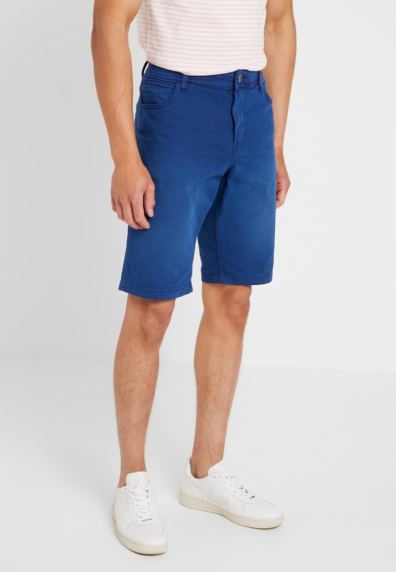 Siniset puuvillashortsit suoralinjaisella leikkauksella, viisi taskua ja nappikiinnitys. Yhdistetty valkoisiin lenkkareihin. Suora pinta, ilman näkyviä kuvioita.