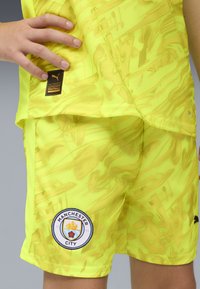 Equipación de fútbol amarilla neón con un patrón texturizado. Incluye el parche del logo de Manchester City y la marca Puma en negro en los pantalones.