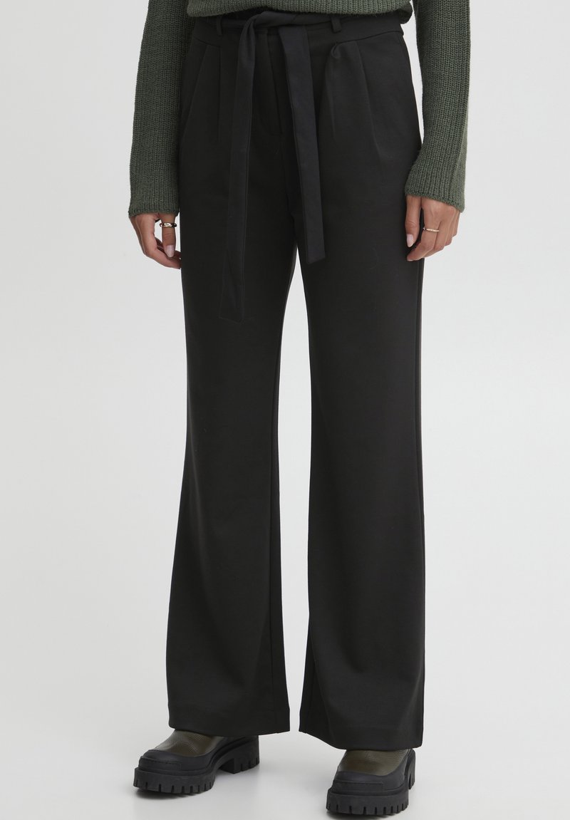 Pantalons larges noirs en tissu doux avec une ceinture à nouer assortie. Présente des plis à l'avant, associés à des bottes noires robustes.