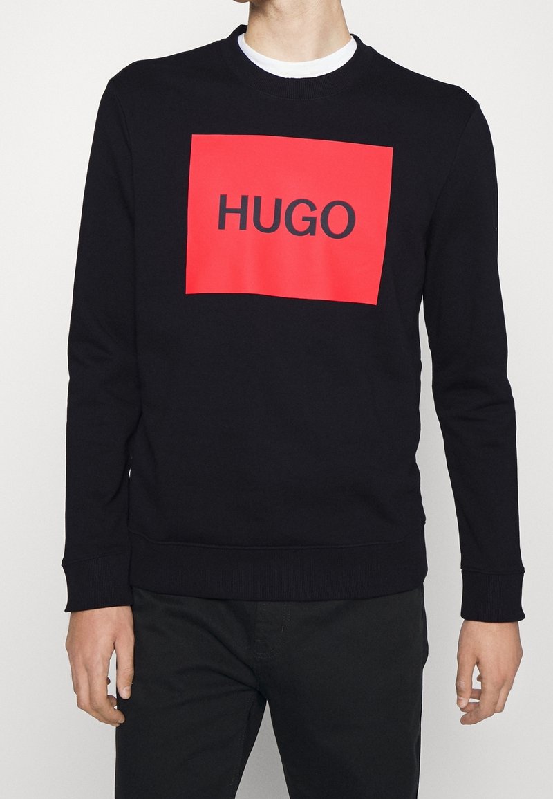 Homme portant un sweat noir avec un grand carré rouge et le mot "HUGO" en lettres noires sur la poitrine.