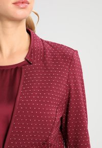 Burgundzka marynarka z teksturowanej tkaniny, zdobiona kropeczkowym wzorem, z kołnierzem mandaryńskim i gładką podszewką.