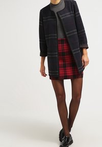 Marineblauwe geruite jas met grijze en blauwe accenten, gecombineerd met een rood-zwarte geruite rok. Model draagt zwarte lakleren schoenen en panty's.