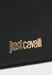 Juodas tekstūruotas rankinės maišas su išryškintu auksiniu „Just Cavalli“ logotipu priekyje. Lygus paviršius su minimalistiniu dizainu.