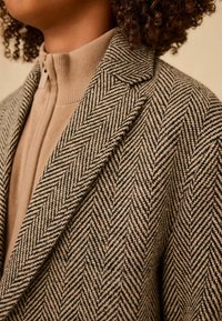 Beige blazer med hægame mønster og struktureret krave, med tekstureret stof og kontraststikninger. Båret over en bluse med høj krave.