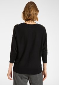 Czarny sweter z rękawami do trzech ćwierci, teksturowana tkanina oraz złote nitowane akcenty wzdłuż ramion. Żebrowany dół i mankiety.