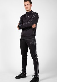 Zwart sportieve trainingspak met witte zijstrepen, met ritszakken en een getailleerd ontwerp. Model draagt zwarte sneakers.