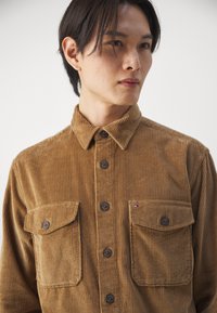 Brown corduroy shirt with a button-down collar, featuring two chest pockets and dark buttons. Subtle ribbed texture and visible stitching details.