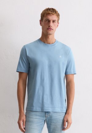 Blonder Mann in hellblauem Kurzarm-T-Shirt und ausgewaschenen blauen Jeans steht vor einer schlichten weißen Wand.
