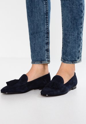 Navy suede loafers with a small heel, featuring a fringe detail at the front. Paired with fitted, lightly distressed blue jeans.