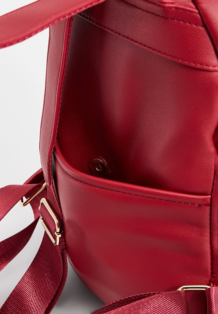 Mochila de cuero rojo con textura suave, herrajes en tono dorado y un bolsillo interior para almacenamiento. Incluye correas de tela ajustables.