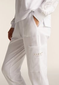 Pantalones de chándal blancos hechos de una tela suave, con un bolsillo lateral que presenta un logo dorado de "FREDDY training" y puños acanalados.