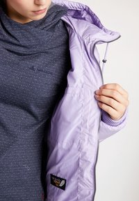 Personne portant un haut bleu foncé à pois, tenant ouverte une veste violet clair avec une étiquette « duvet haute qualité 700 » à l'intérieur.
