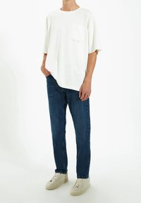 Camiseta blanca de algodón con mangas cortas y un bolsillo en el pecho, combinada con vaqueros de mezclilla azul oscuro y zapatillas de color claro.