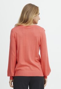 Pull-over en tricot corail avec un col rond, des manches longues et des poignets côtelés, présentant une texture lisse et une coupe décontractée.
