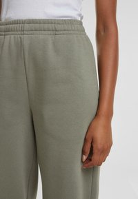 Lysegrønne joggebukser har en elastisk midje og en glatt, myk tekstur. En hånd hviler på stoffet, noe som viser en nærbildevisning.