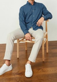Niebiesko-czarna koszula w kratę na guziki, beżowe spodnie w slim fit oraz białe trampki. Gładki materiał i klasyczny krój, siedzący na drewnianym krześle.