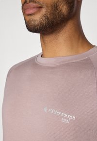 Lys rosa hettegenser med teksturert overflate, med rund hals og logo-detalj på brystet. Myk stoff med minimal søm.