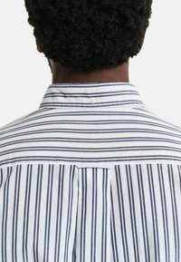 Chemise blanche à rayures verticales bleu marine, col boutonné et un détail de pli au dos. Tissu en coton doux avec une texture lisse.
