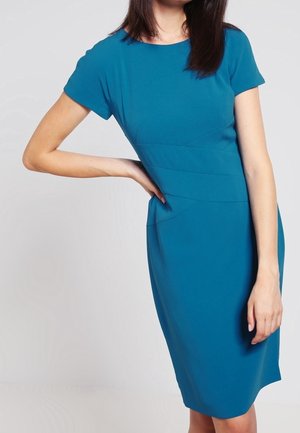 Frau in einem knieumspielenden, kurzärmeligen Kleid in Türkis, steht mit einer Hand in der Hüfte und dem anderen Arm entspannt an der Seite.