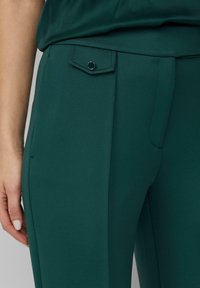 Ciemnozielone, dopasowane spodnie o gładkiej fakturze, z małą klapką kieszeni z przodu i okrągłym ozdobnym guzikiem.