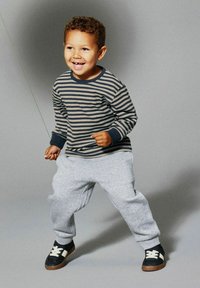 Uśmiechnięty młody chłopiec w paskowanej koszulce z długim rękawem, szarych dresach i trampkach stoi z nogami rozstawionymi na szarym tle.