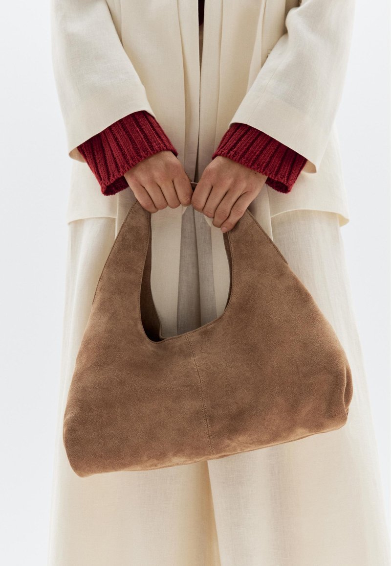 Borsa in suede di colore marrone chiaro, con forma arrotondata e manico superiore. Tenuta da mani che indossano una manica rossa a maglia, appoggiata su un indumento color crema.