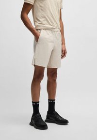 Pantalones cortos ligeros beige con cintura elástica, bolsillos laterales y detalles de cremallera. Se combinan con zapatillas negras y calcetines con texto de logo.